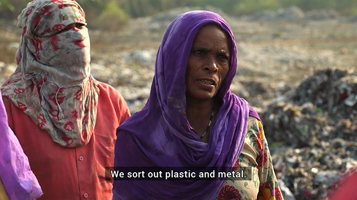 Women in headscarves standing in landfill with closed caption, "We sort out plastic and metal."