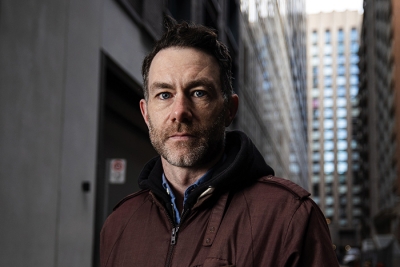 Brendan Kennedy, a man with short hair and a trimmed beard, stands in an urban alleyway, wearing a brown jacket over a hoodie.